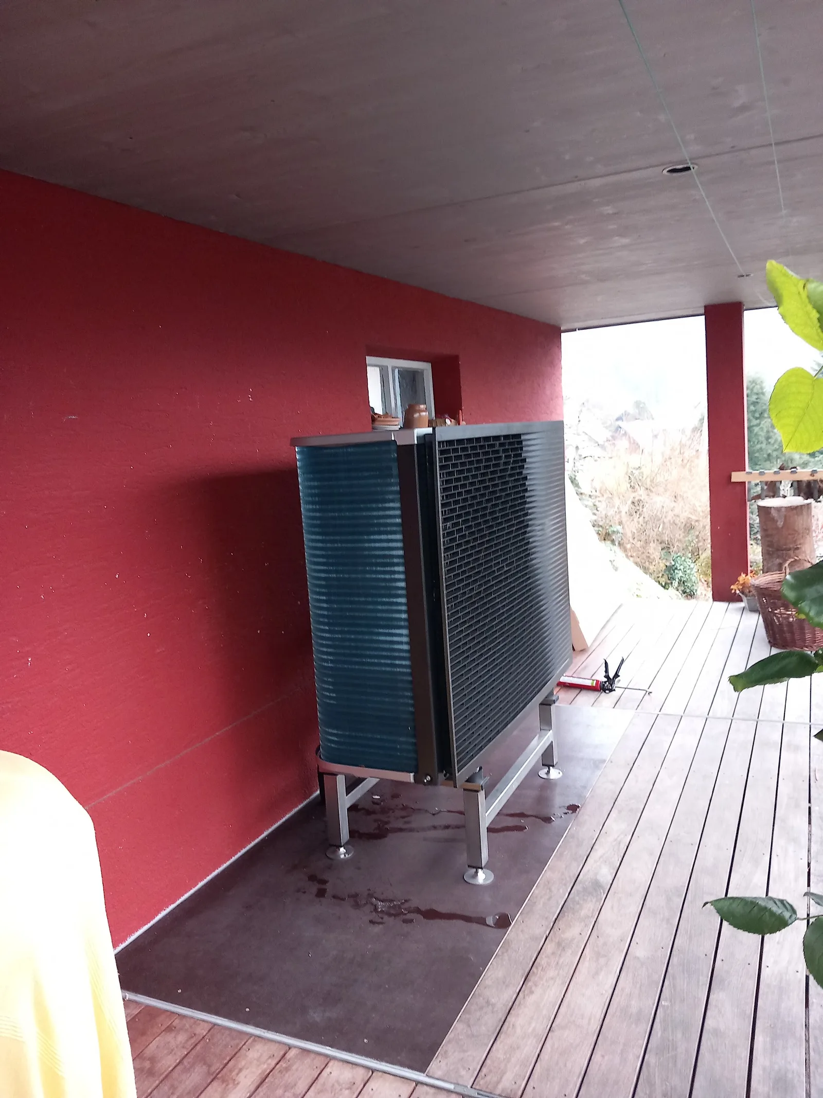 Luft-Wärmepumpe unter einem Vordach an einer roten Hausfassade, davor eine Holzterrasse